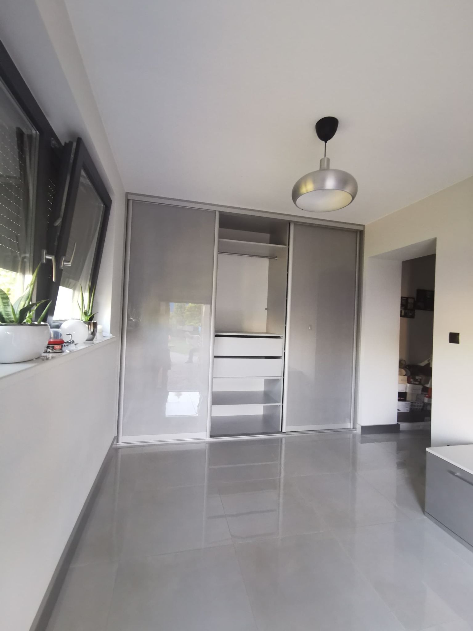 Nowoczesna szafa wnękowa z przesuwnymi, szklanymi drzwiami w szarym odcieniu, widoczna w minimalistycznym wnętrzu z błyszczącą, szarą podłogą i roślinami doniczkowymi na parapecie okiennym.