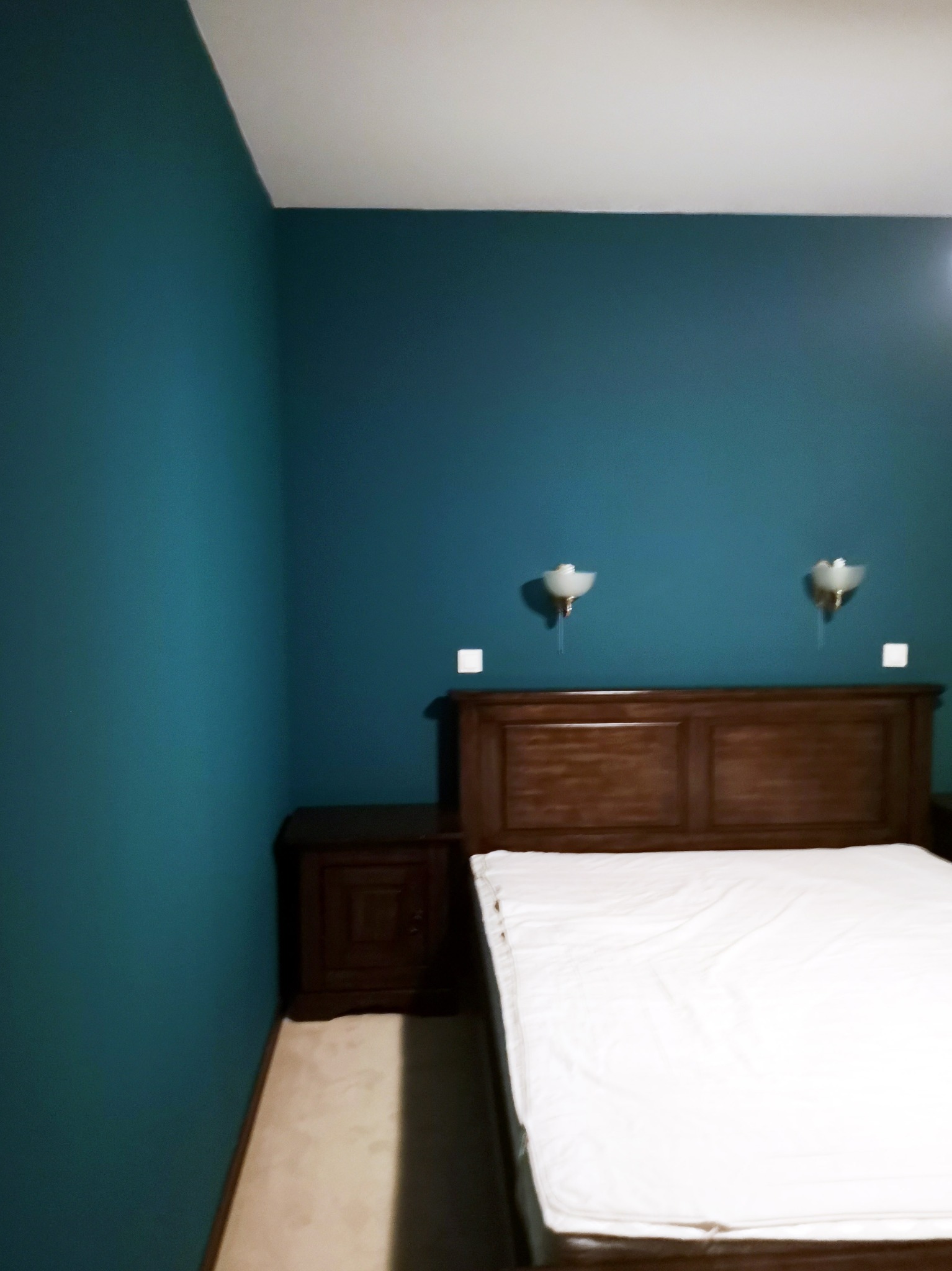Sypialnia z łóżkiem z ciemnego drewna, turkusowymi ścianami i lampkami nocnymi, widok z boku.