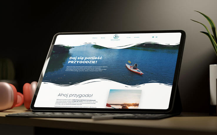 Tablet prezentuje stronę internetową z motywem kajakarstwa, hasłem 'Daj się ponieść przygodzie!' i zdjęciem osoby płynącej kajakiem po jeziorze. Minimalistyczny design.