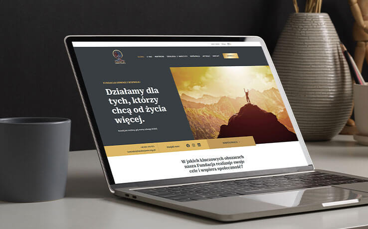 Laptop prezentuje stronę internetową fundacji z motywem górskim i hasłem 'Działamy dla tych, którzy chcą od życia więcej'. Stonowana kolorystyka, minimalistyczny design.