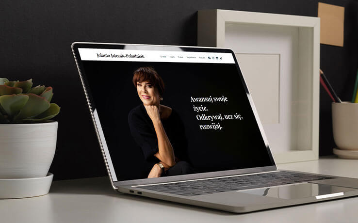Laptop wyświetla stronę internetową z portretem kobiety i hasłem 'Awansuj swoje życie. Odkrywaj, ucz się, rozwijaj.', obok roślina w doniczce i ramka na zdjęcia.