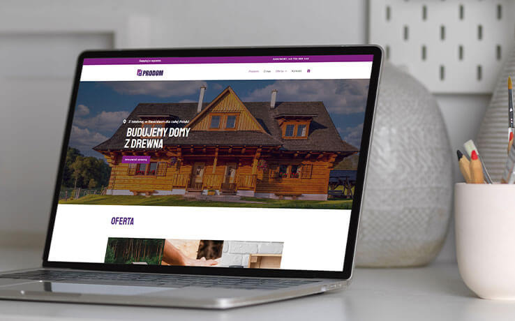 Laptop wyświetla stronę internetową firmy budującej domy z drewna. W tle wazon i przybory do pisania. Minimalistyczne biurko, stonowana kolorystyka.