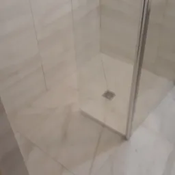 Wykończona łazienka z kabiną prysznicową z przezroczystego szkła i płytkami w odcieniach beżu, widoczny fragment umywalki na postumencie.