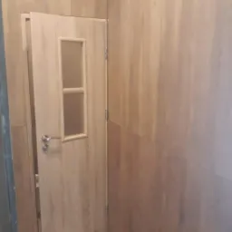 Drewnopodobne panele ścienne w odcieniach brązu i beżu, z otwartymi drzwiami z przeszkleniem i czterema otworami wentylacyjnymi u dołu.