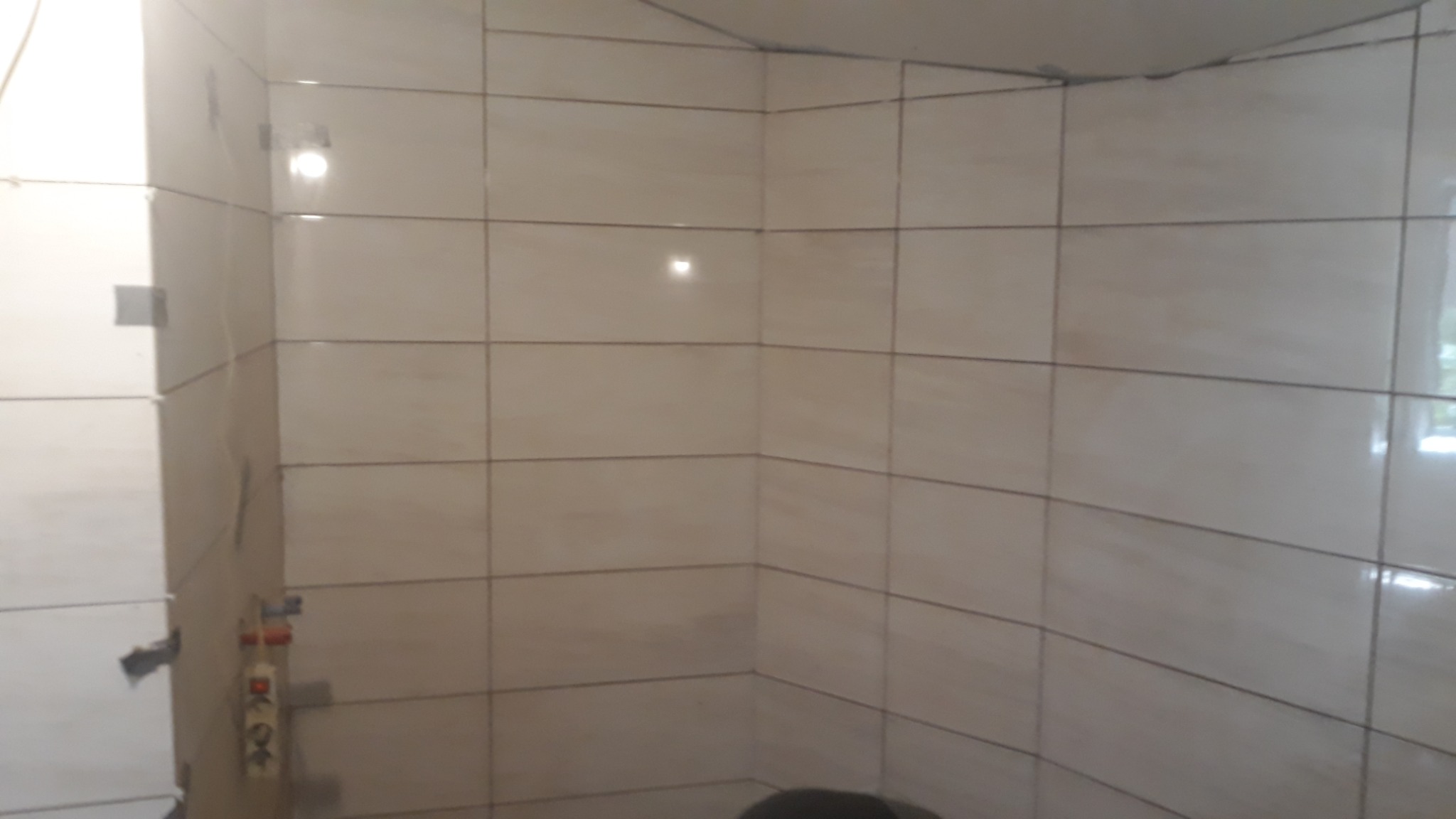 Ściana łazienki wyłożona jasnymi, beżowymi płytkami ceramicznymi, widoczne fugi w kolorze grafitowym, fragment instalacji wodnej z widocznymi rurami i przyłączami, narożnik pomieszczenia.