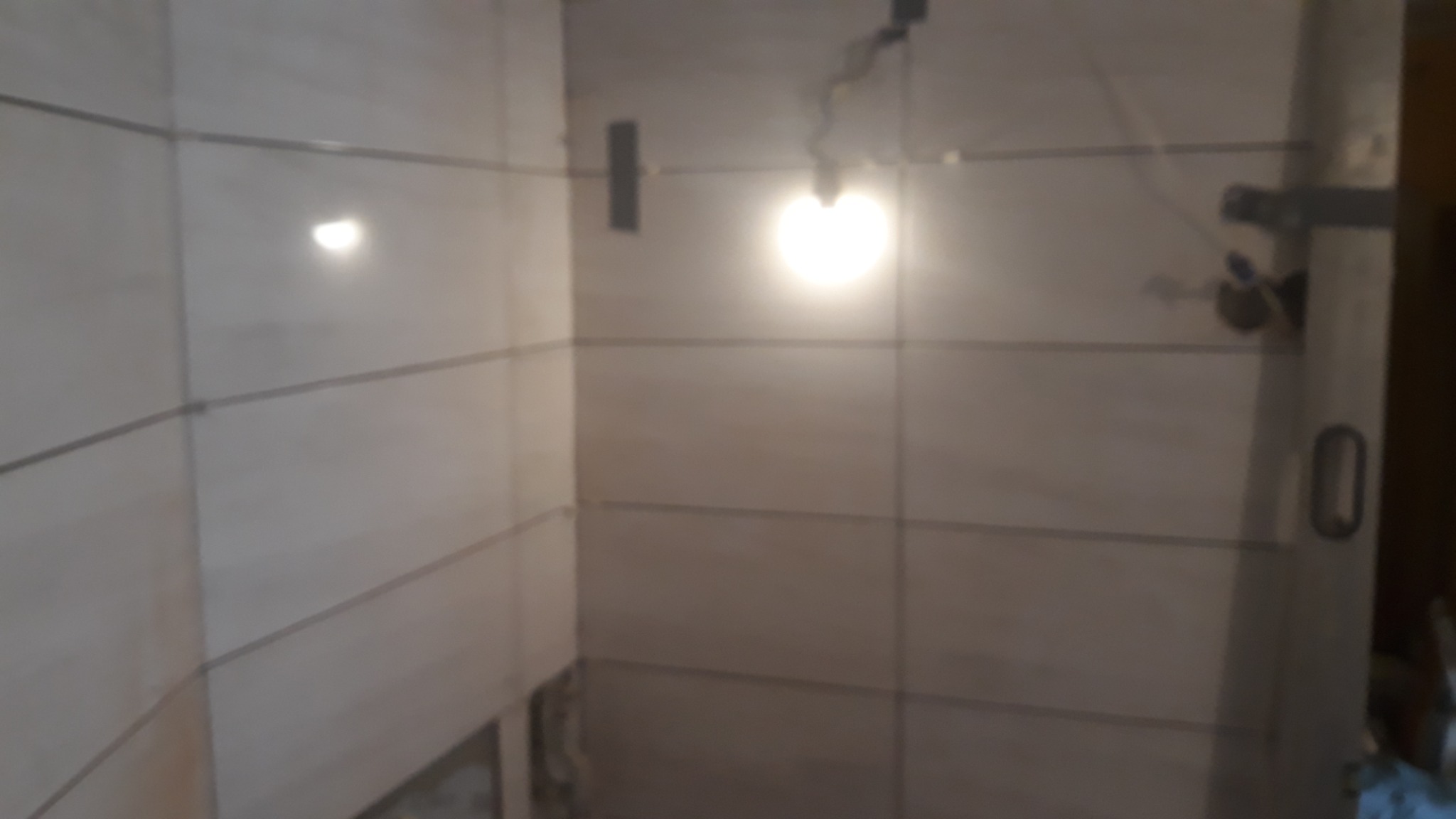 Ściany łazienki wyłożone jasnymi płytkami ceramicznymi z ciemnymi fugami, widoczne niedokończone prace przy podłodze i oświetlenie budowlane.