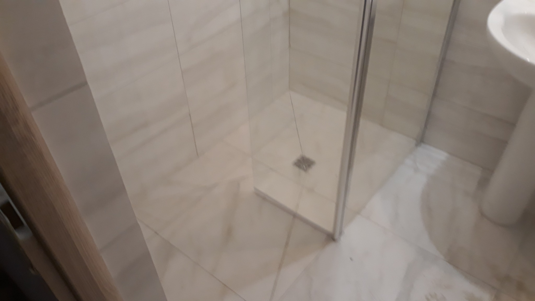 Wykończona łazienka z kabiną prysznicową z przezroczystego szkła i płytkami w odcieniach beżu, widoczny fragment umywalki na postumencie.