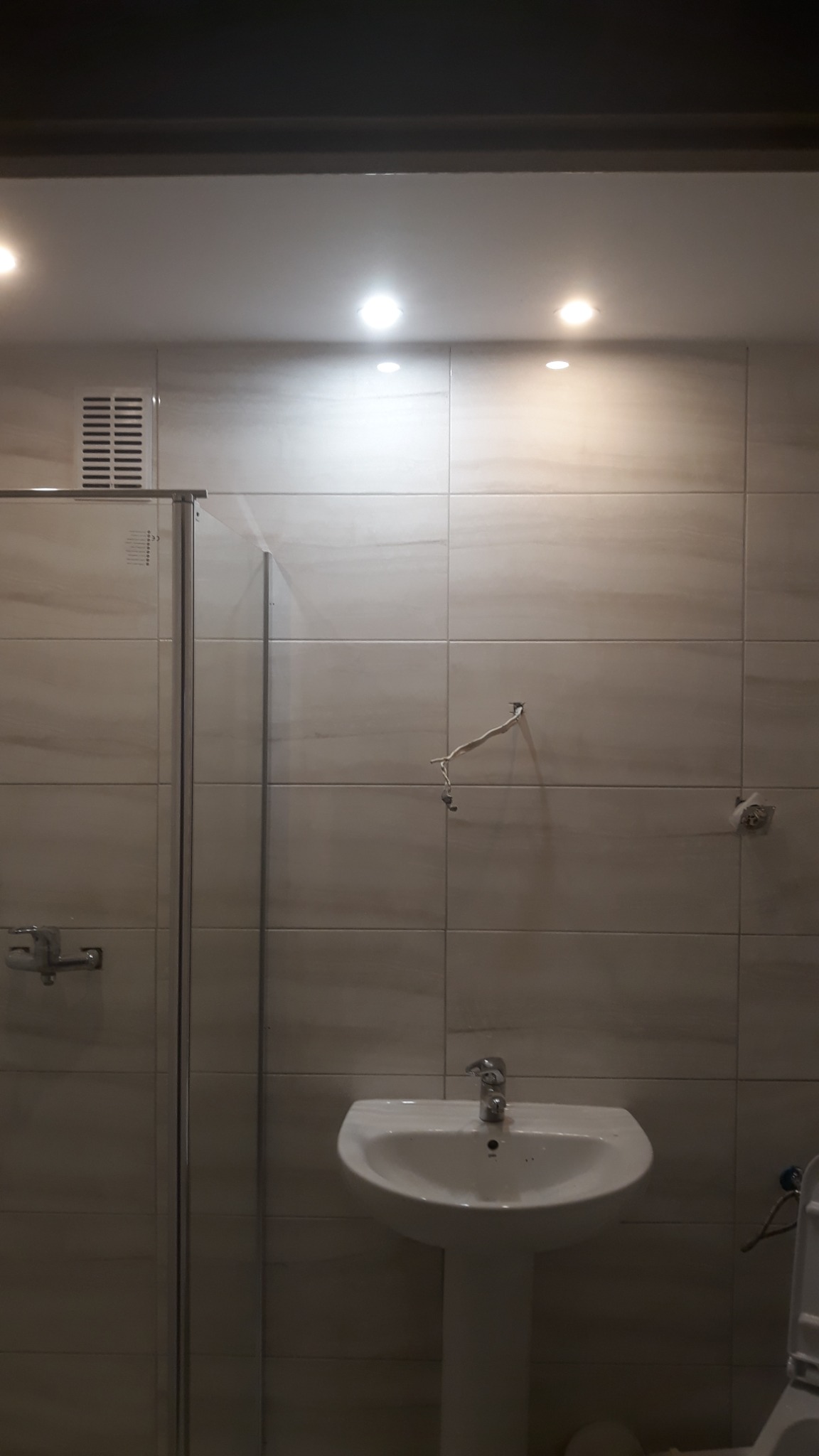 Łazienka w trakcie remontu: widoczne płytki ścienne w odcieniach beżu, kabina prysznicowa z chromowanymi elementami, umywalka na postumencie oraz wystające kable ze ściany, sugerujące prace...