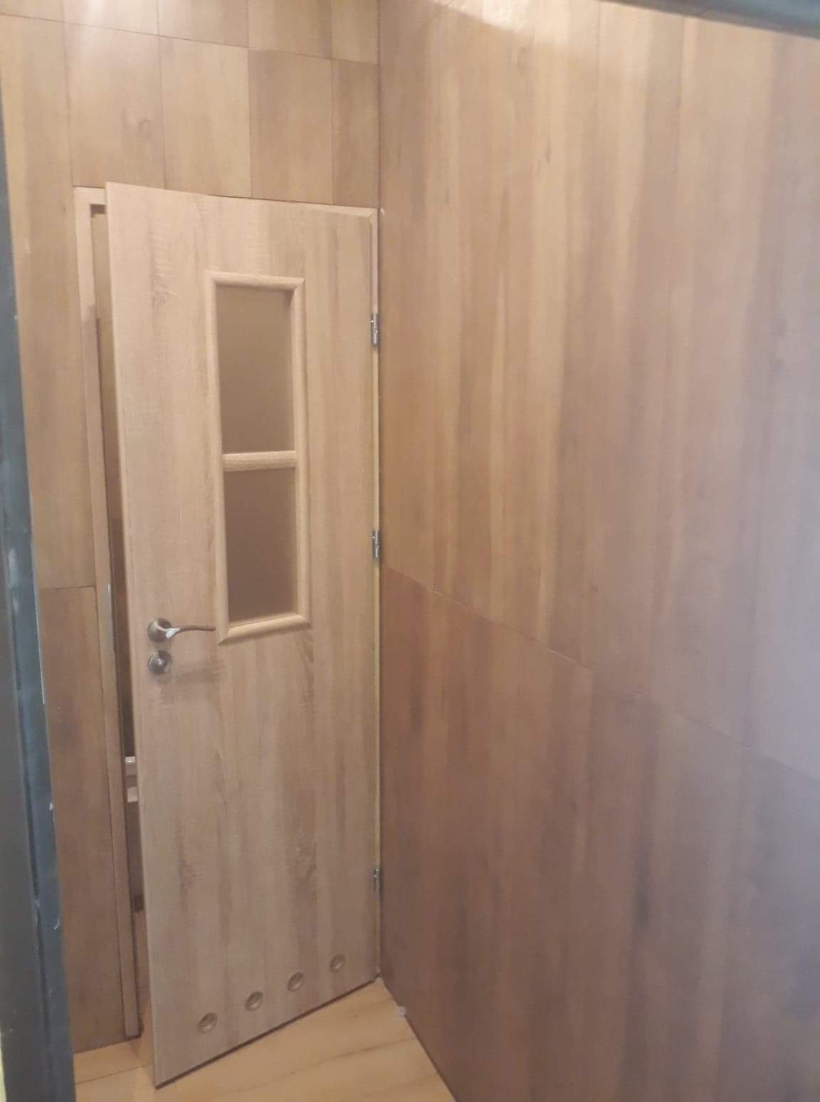 Drewnopodobne panele ścienne w odcieniach brązu i beżu, z otwartymi drzwiami z przeszkleniem i czterema otworami wentylacyjnymi u dołu.