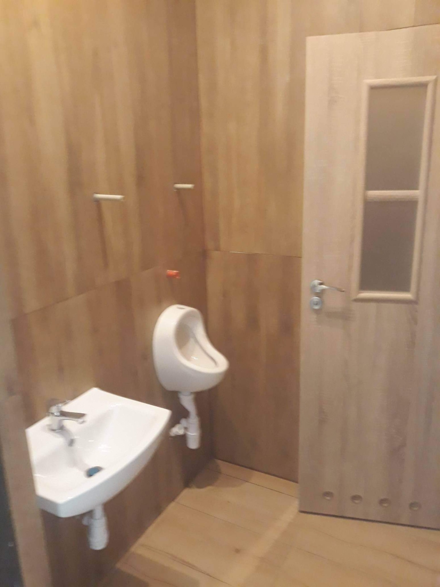 Ukończona toaleta z umywalką, pisuar i drewnopodobnymi płytkami na ścianach oraz drewnianymi drzwiami z szybką.