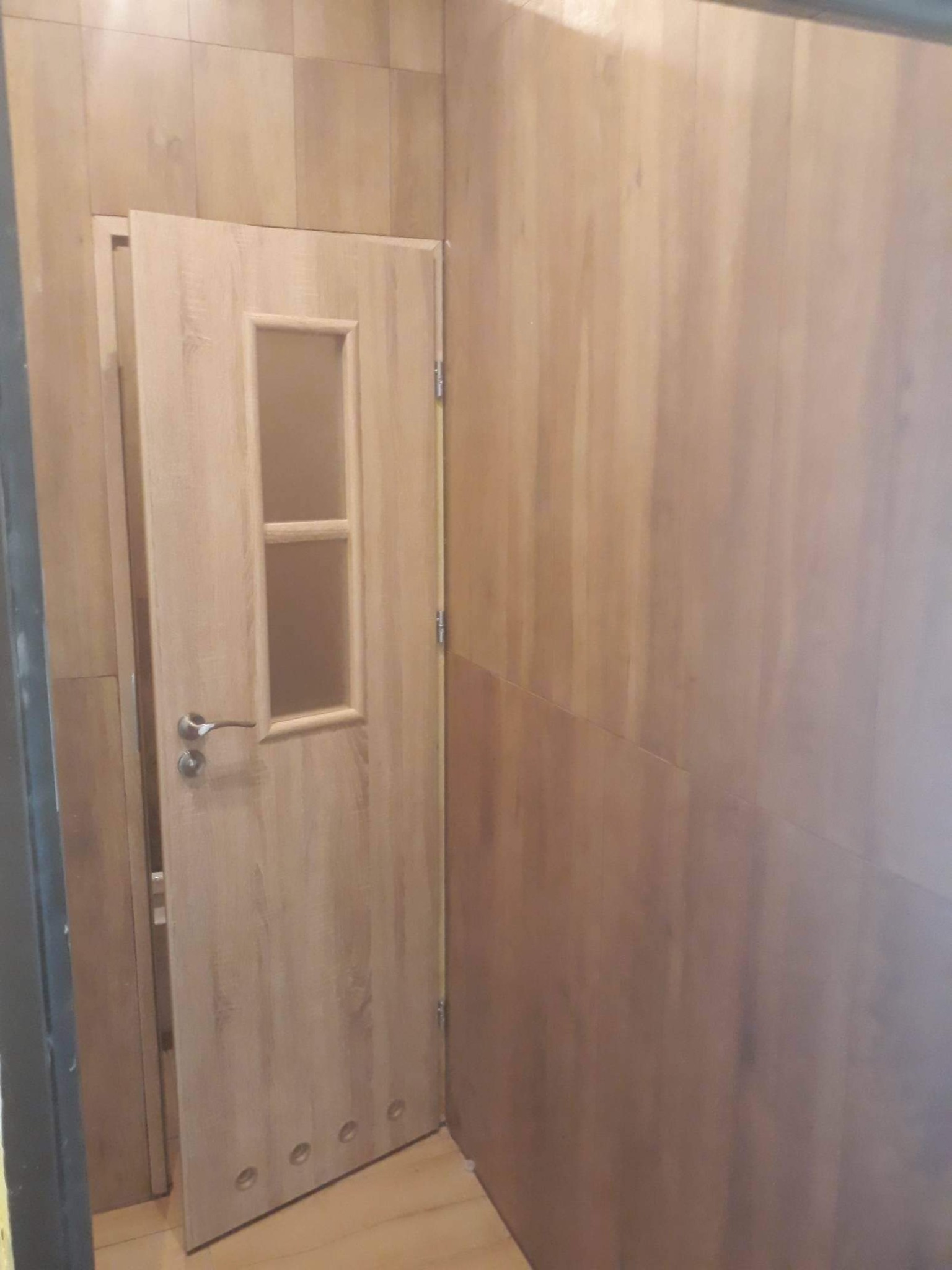 Otwarta drewniana drzwi wewnętrzne z szybą, dopasowane do drewnianej okładziny ściennej, widoczne cztery otwory wentylacyjne w dolnej części skrzydła.