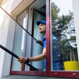 F.M CORP. SPÓŁKA Z OGRANICZONĄ ODPOWIEDZIALNOŚCIĄ - Montaż moskitiery okiennej przez pracownika w niebieskim uniformie. Widoczny fragment elewacji budynku i żółte wiadro.