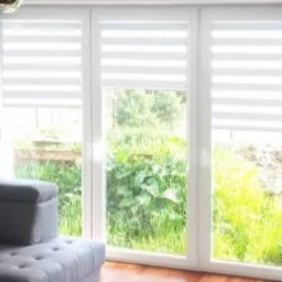 F.M CORP. SPÓŁKA Z OGRANICZONĄ ODPOWIEDZIALNOŚCIĄ - Okno z trzema skrzydłami, przesłonięte roletami dzień-noc w biało-szare pasy, z widokiem na zieleń. Szara zasłona i fragment sofy w tle.