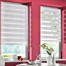 F.M CORP. SPÓŁKA Z OGRANICZONĄ ODPOWIEDZIALNOŚCIĄ - Dwa okna z roletami dzień noc w białej kolorystyce. Na parapecie wazon z kwiatami, dzbanek i owoce. Intensywny różowy kolor ścian.