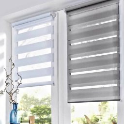 F.M CORP. SPÓŁKA Z OGRANICZONĄ ODPOWIEDZIALNOŚCIĄ - Dwa okna z zamontowanymi roletami dzień noc w kolorze białym i szarym. Na parapecie dekoracje: wazon, książki, kieliszek i ozdobne pióro.