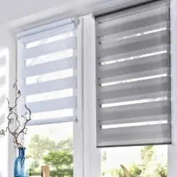 Dwa okna z zamontowanymi roletami dzień noc w kolorze białym i szarym. Na parapecie dekoracje: wazon, książki, kieliszek i ozdobne pióro.