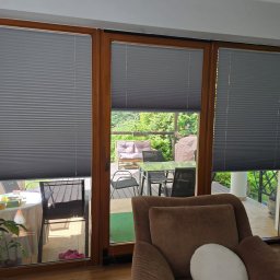 F.M CORP. SPÓŁKA Z OGRANICZONĄ ODPOWIEDZIALNOŚCIĄ - Szare plisy okienne zamontowane w brązowych ramach okiennych, widok na taras z meblami ogrodowymi. Widoczne fałdy i struktura materiału plis.