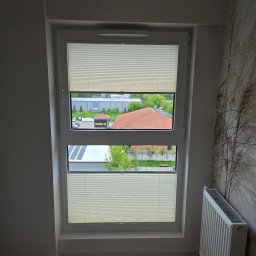 F.M CORP. SPÓŁKA Z OGRANICZONĄ ODPOWIEDZIALNOŚCIĄ - Beżowe plisy okienne zamontowane w oknie dwudzielnym, z widokiem na budynek sklepu Netto i dom z czerwoną dachówką. Grzejnik pod oknem.