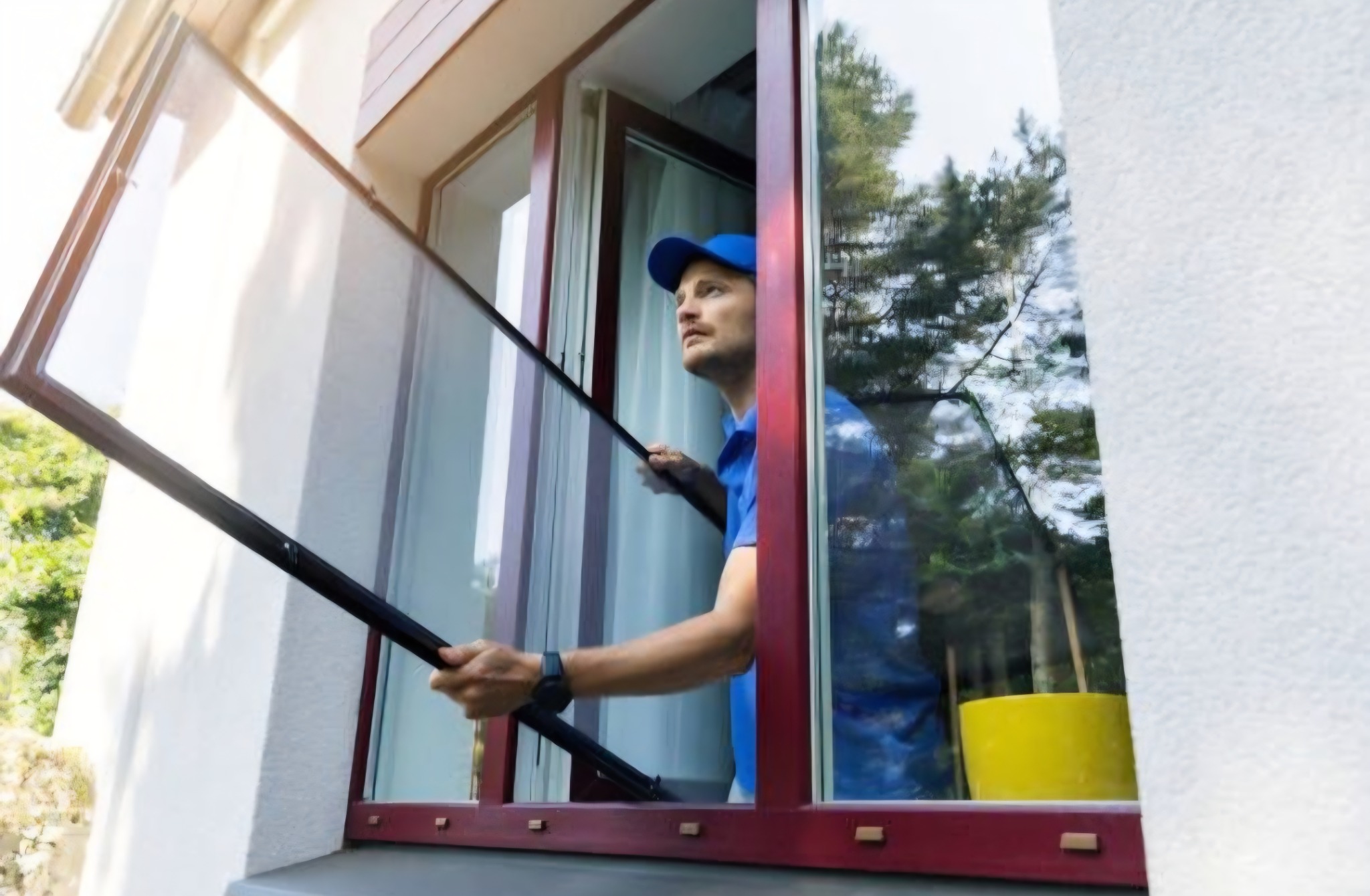 Montaż moskitiery okiennej przez pracownika w niebieskim uniformie. Widoczny fragment elewacji budynku i żółte wiadro.