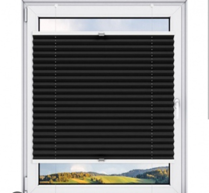 Czarne plisy okienne zamontowane w białym oknie PCV, widok na krajobraz za oknem. Plisa częściowo zasłonięta, widoczne mocowania.