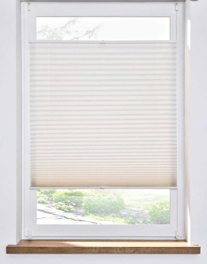 Jasne plisy okienne, częściowo zasłonięte, z widokiem na zieleń za oknem. Ustawione w oknie z drewnianym parapetem. Stonowana kolorystyka.