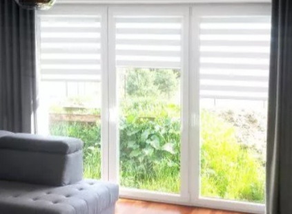 Okno z trzema skrzydłami, przesłonięte roletami dzień-noc w biało-szare pasy, z widokiem na zieleń. Szara zasłona i fragment sofy w tle.