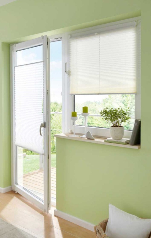 Jasne plisy okienne zamontowane w oknie i na drzwiach balkonowych, z widokiem na zieleń za oknem. Dekoracja okienna w nowoczesnym, minimalistycznym stylu.