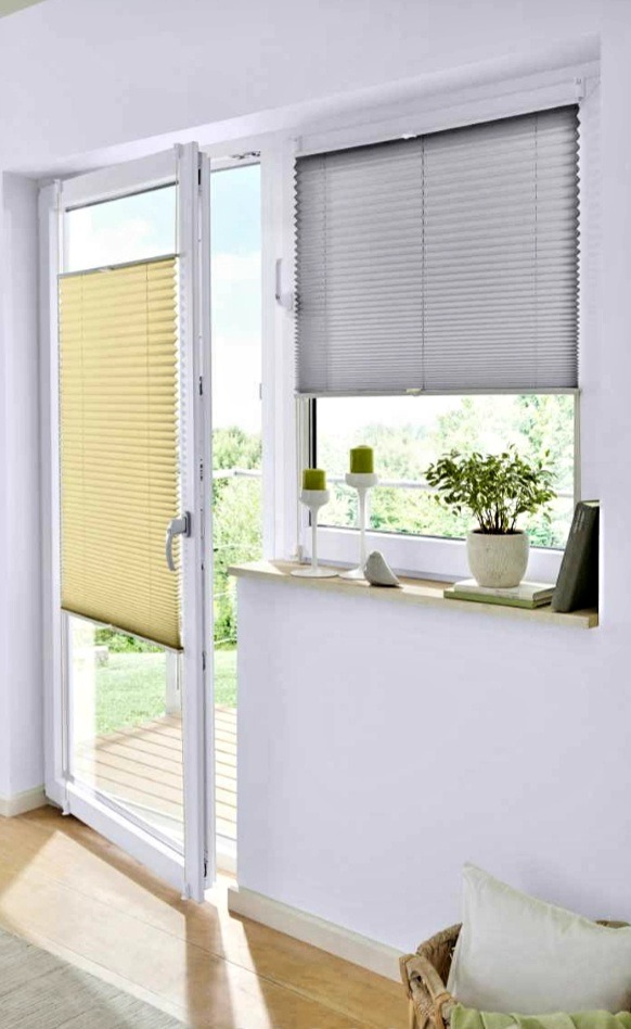 Beżowa i szara plisa okienna na oknie balkonowym, z dekoracjami na parapecie i widokiem na zieleń. Nowoczesny design, jasne wnętrze.