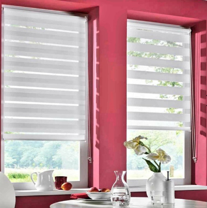 Dwa okna z roletami dzień noc w białej kolorystyce. Na parapecie wazon z kwiatami, dzbanek i owoce. Intensywny różowy kolor ścian.