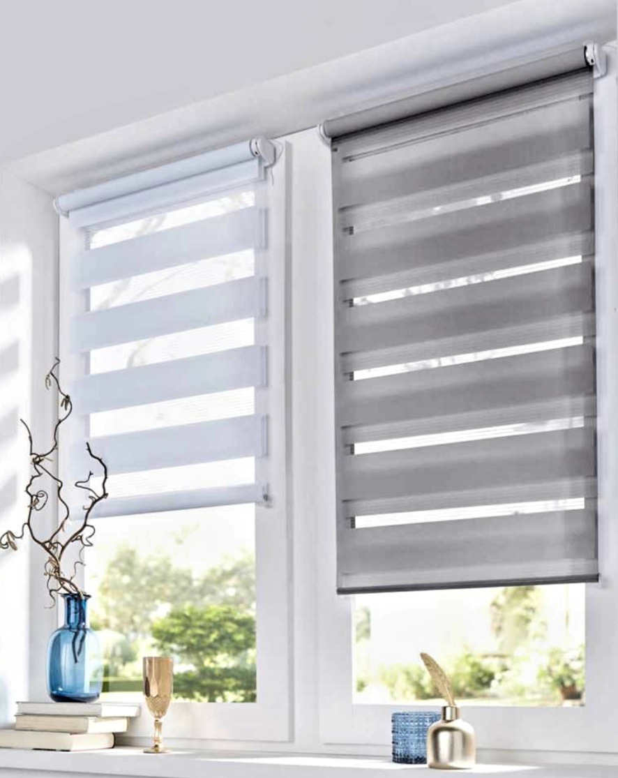 Dwa okna z zamontowanymi roletami dzień noc w kolorze białym i szarym. Na parapecie dekoracje: wazon, książki, kieliszek i ozdobne pióro.