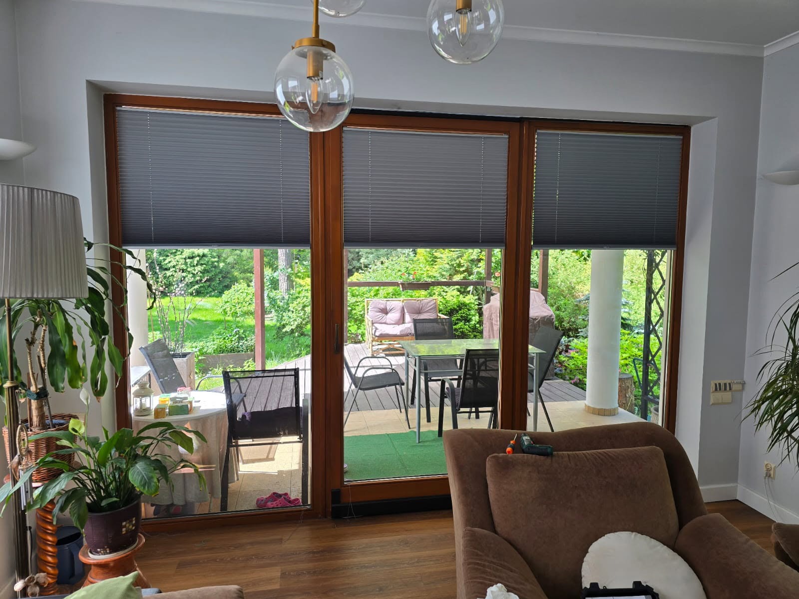 Szare plisy okienne zamontowane w dużym oknie tarasowym z drewnianymi ramami. Widok na zielony ogród i taras z meblami wypoczynkowymi. Wnętrze domu z roślinami.