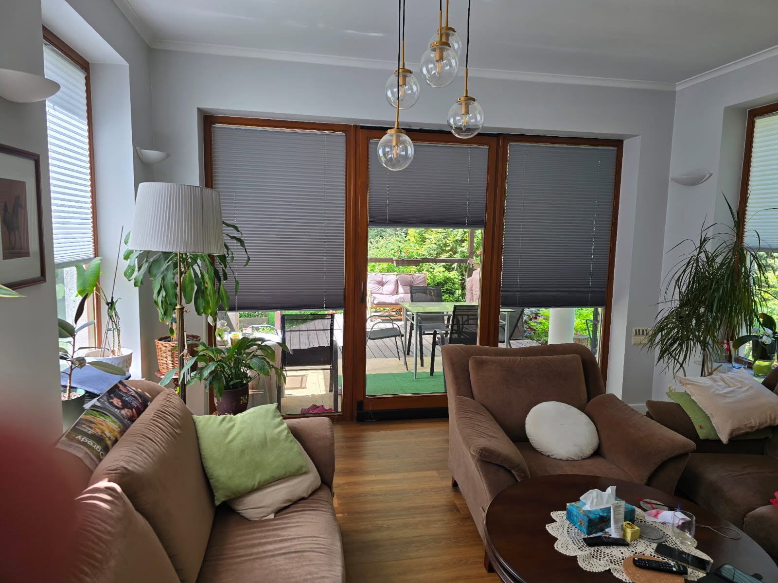 Szare plisy okienne zamontowane w oknach tarasowych drewnianej ramy. Wnętrze salonu z brązowymi meblami i widokiem na zielony ogród.