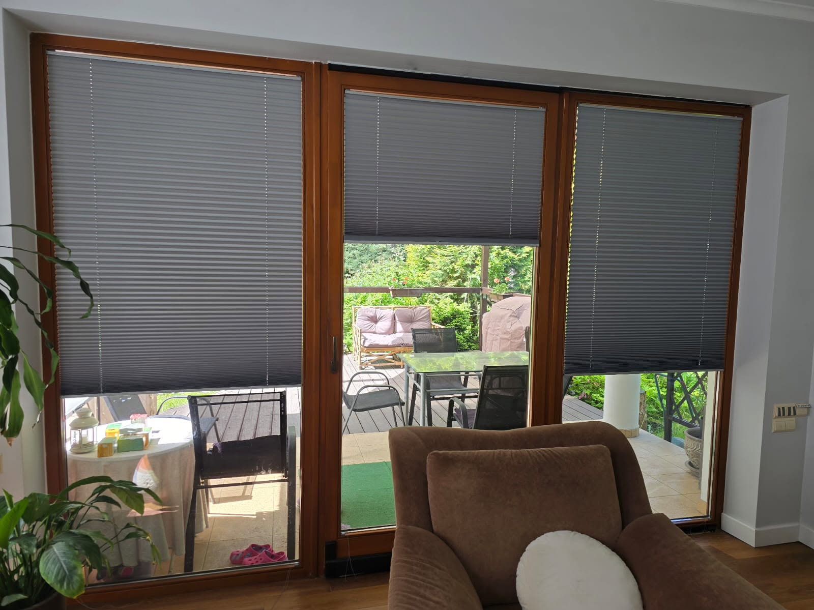 Szare plisy okienne zamontowane w brązowych ramach okiennych, widok na taras z meblami ogrodowymi. Widoczne fałdy i struktura materiału plis.