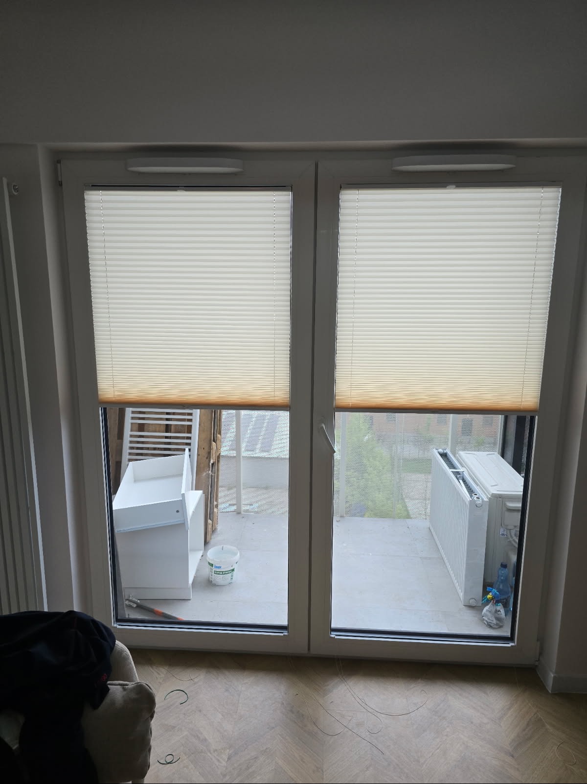 Białe plisy zamontowane w dwuskrzydłowym oknie balkonowym. Widok na balkon z widocznym grzejnikiem i elementami wyposażenia. Delikatne światło dzienne.