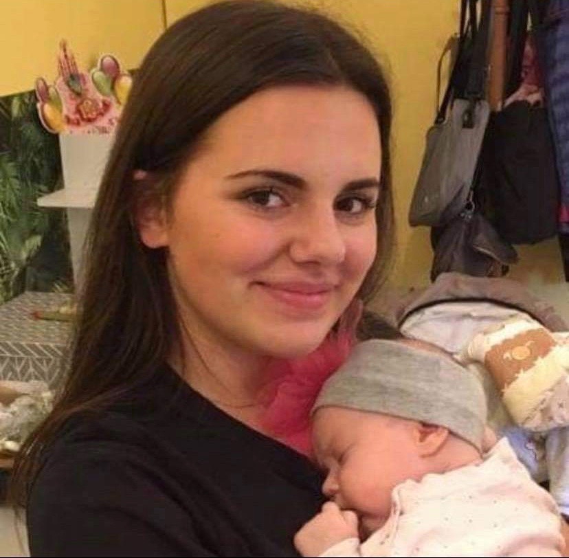 Uśmiechnięta młoda kobieta trzyma śpiące niemowlę w szarej czapeczce, różowa ozdoba na szyi, w tle wieszak z ubraniami i dekoracja urodzinowa.