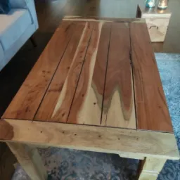 Stolik kawowy z blatem z desek z wyraźnym usłojeniem, nogi z jasnego drewna, na tle dywanu z wzorem i fragmentem sofy.