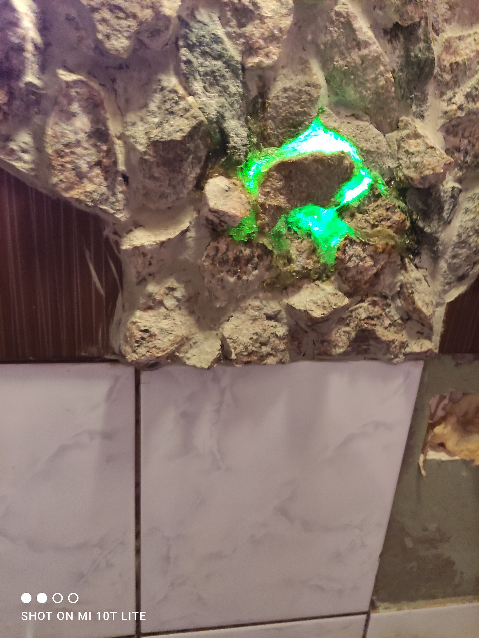 Fragment dekoracyjnej ściany z kamieni z zielonym podświetleniem, połączonej z białymi płytkami imitującymi marmur i brązową boazerią.