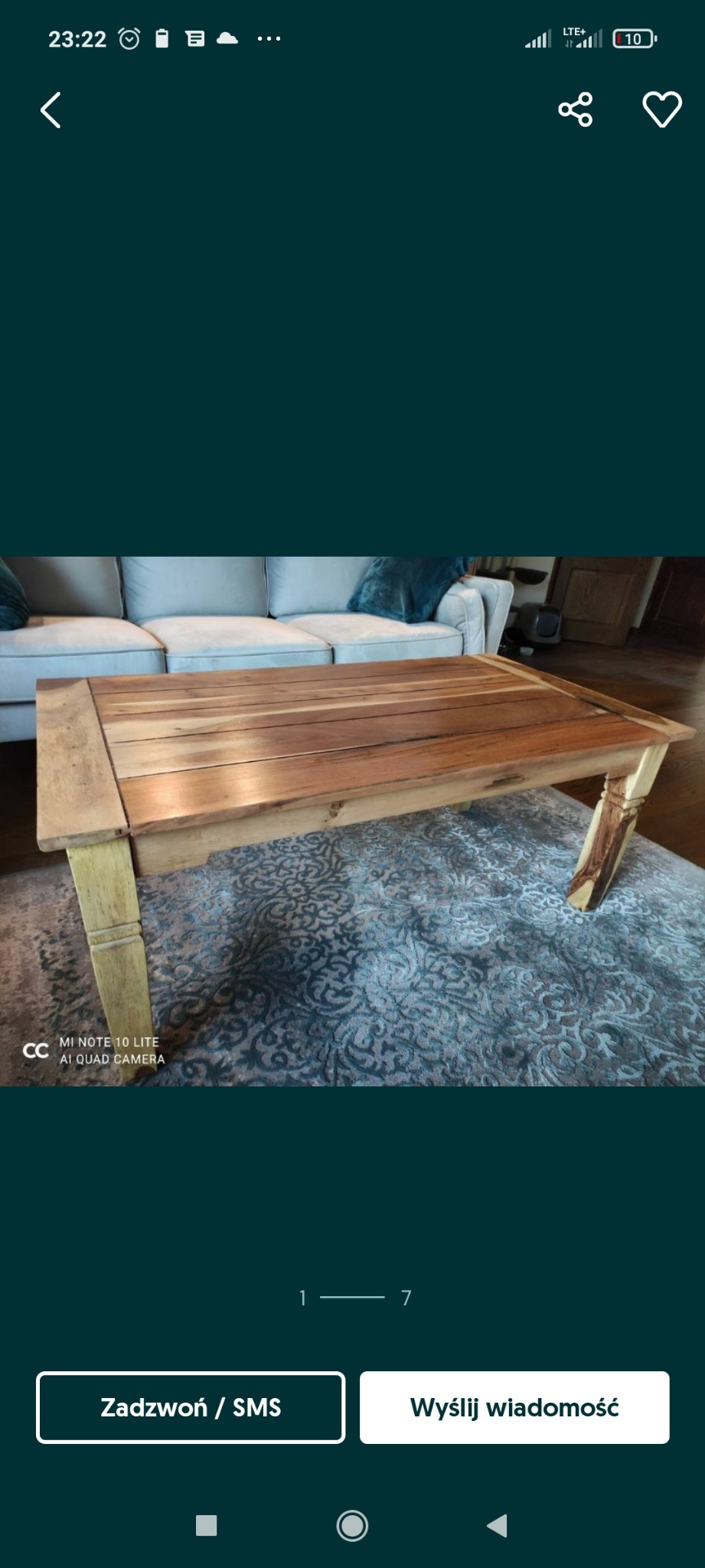 Drewniany stolik kawowy z jasnymi nogami na wzorzystym dywanie, w tle jasna sofa.