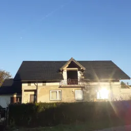 Dom jednorodzinny z nowym, czarnym dachem z dachówki ceramicznej, w trakcie budowy lukarny, widoczne rusztowanie na balkonie, elewacja częściowo otynkowana, w tle błękitne niebo i smuga...