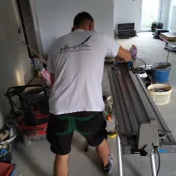 Pracownik w koszulce z logo firmy AG Mont przycina płytki na elektrycznej maszynce w trakcie prac wykończeniowych w pomieszczeniu z oknem.