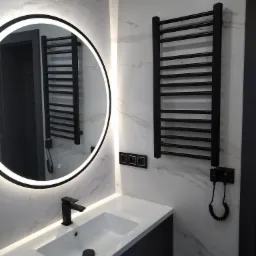Nowoczesna łazienka z marmurową ścianą, okrągłym lustrem z podświetleniem LED, czarną armaturą i grzejnikiem drabinkowym.