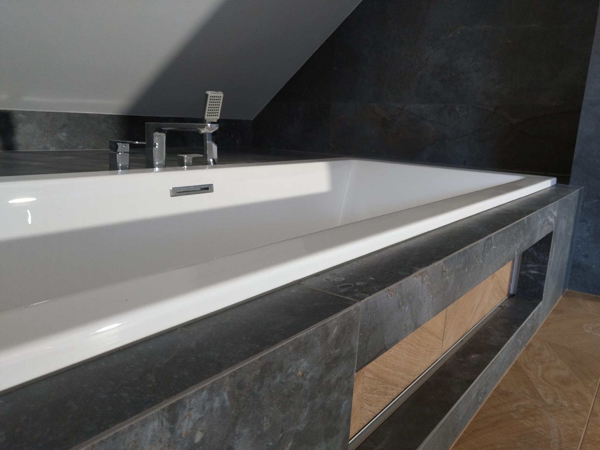 Biała wanna w zabudowie z szarego kamienia w nowoczesnej łazience na poddaszu, z chromowaną armaturą i mozaiką w tle.