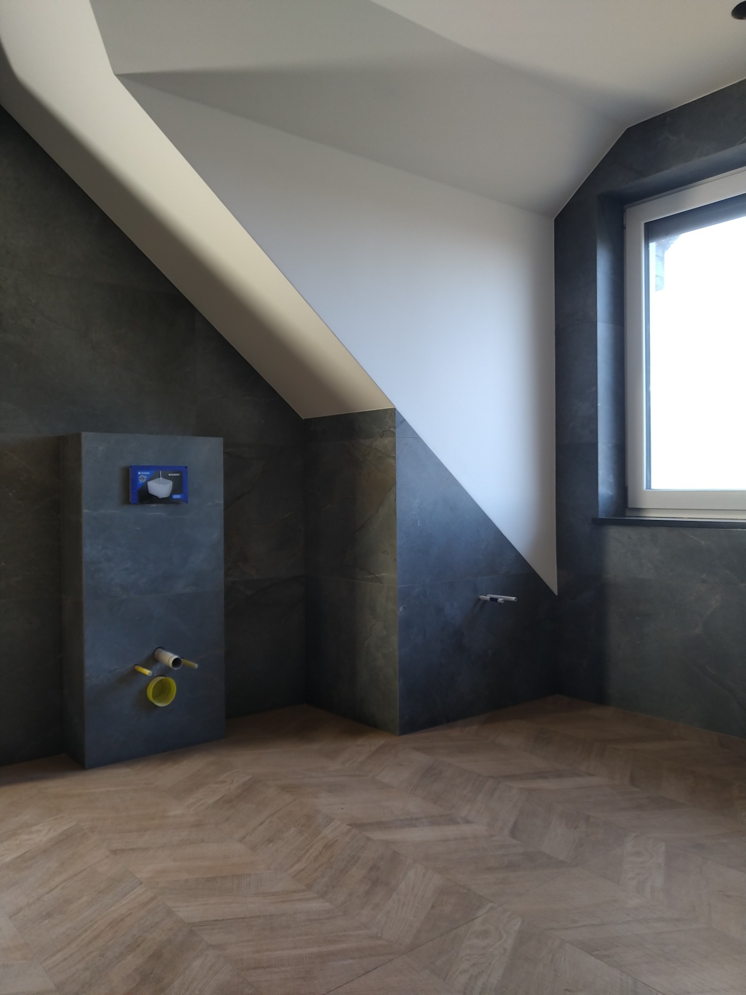 Nowoczesna łazienka na poddaszu z ciemnymi płytkami imitującymi kamień, drewnianą podłogą ułożoną w jodełkę, zabudowaną toaletą podtynkową i oknem w ciemnej ramie.