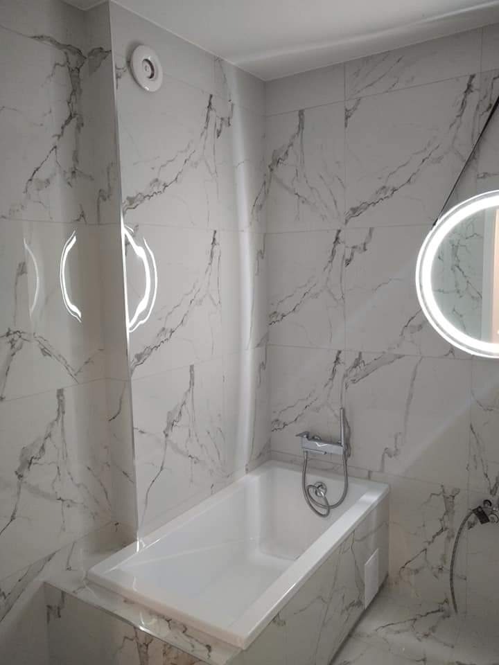 Wykończona łazienka z wanną obłożoną białymi płytkami imitującymi marmur, okrągłe podświetlane lustro i wentylator sufitowy.