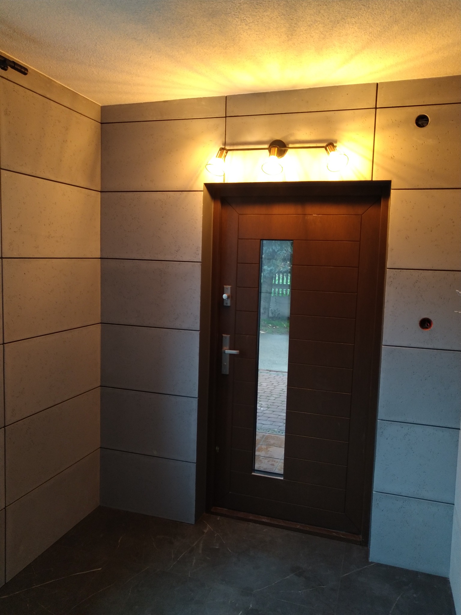 Wnętrze przedpokoju z ciemną, drewnianą stolarką drzwiową z przeszkleniem, wykończone płytami imitującymi beton w szarym odcieniu, oświetlone trzypunktową lampą sufitową.