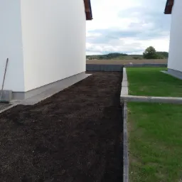 Przygotowany teren pod trawnik między dwoma budynkami, z widoczną świeżo rozłożoną ziemią, obrzeżami z kostki i fragmentem trawnika z tyłu.