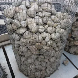 Cylindryczny kosz gabionowy wypełniony otoczakami w odcieniach szarości i beżu, ustawiony na betonowej płycie, z widocznymi elementami konstrukcji z metalowej siatki.