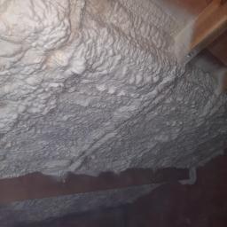 Warming Pur Piotr Jasinski - Szczegółowe ujęcie grubej warstwy białej pianki izolacyjnej, nierównomiernie nałożonej na drewniane elementy konstrukcyjne poddasza, z widocznymi nieregularnościami i pęcherzykami powietrza...