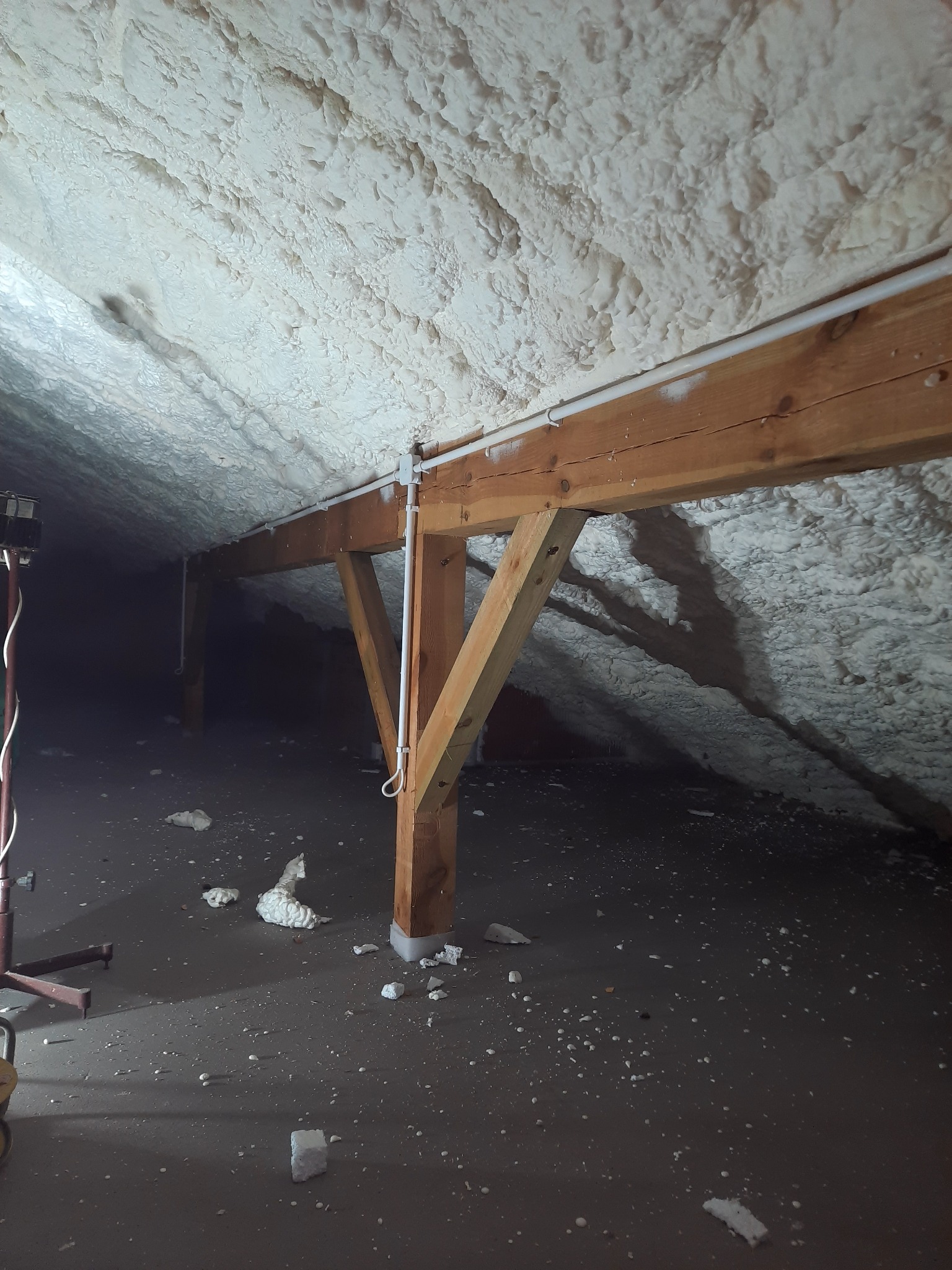 Poddasze z drewnianą konstrukcją pokryte grubą warstwą białej pianki izolacyjnej, widoczne fragmenty odciętej pianki na podłodze, oświetlone lampą roboczą.