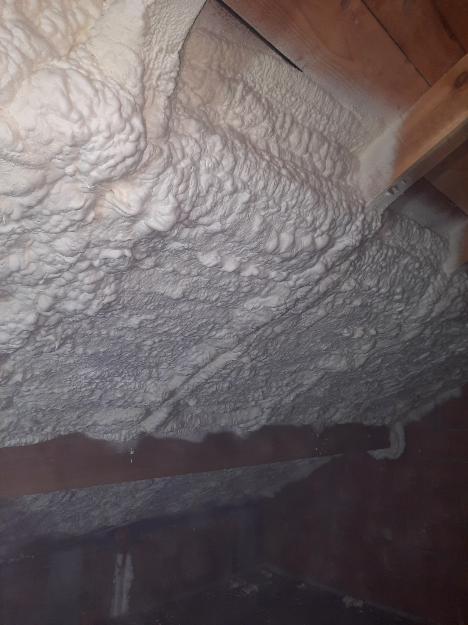 Szczegółowe ujęcie nierównej powierzchni białej pianki izolacyjnej, pokrywającej drewniane elementy konstrukcyjne poddasza.